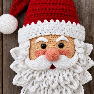 May include: A crocheted Santa Claus face decoration featuring a red hat with a white pom-pom, a white crocheted brim, a peach-colored face with rosy cheeks, black bead eyes, and a textured white beard and mustache made of yarn.