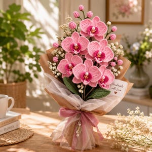 May include: A bouquet of crocheted pink orchids with dark pink centers and small white pearl accents. The flowers are arranged with white filler flowers and green leaves, wrapped in brown paper and white tulle with a pink ribbon and a tag reading Handmade with Love.