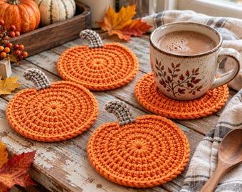 May include: Three round, orange crochet coasters shaped like pumpkins, each featuring a small, textured gray stem at the top. One coaster supports a speckled ceramic mug filled with a brown beverage, while the others rest on a wooden surface.