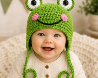 May include: A bright green crocheted hat featuring a frog design with two large eyes on top, each with a white border and a black button pupil. The hat includes two pink circular cheeks and a black stitched smile, with long dangling earflaps.