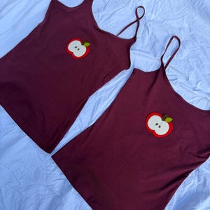 Op de afbeelding: Twee donkerbordeauxrode hemdjes met dunne spaghettibandjes. Elk topje is voorzien van een gecentreerde, geborduurde appelpatch op de borst. Het appelontwerp bestaat uit een rode omtrek, een wit midden en een klein bruin steeltje met een groen blaadje.
