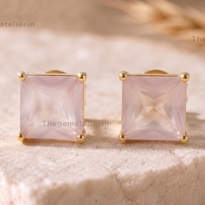 Peut inclure: Une paire de boucles d'oreilles à clous avec une pierre précieuse rose pâle de forme carrée montée sur métal doré. Chaque boucle présente une pierre à facettes avec un sertissage à quatre griffes. Le texte Thegematelierin est visible.