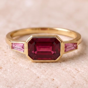 May include: A gold ring featuring a central emerald-cut deep red gemstone flanked by two smaller, tapered baguette-cut pink gemstones. The metal band is smooth and polished, holding the stones in a secure bezel setting with clean, geometric lines.