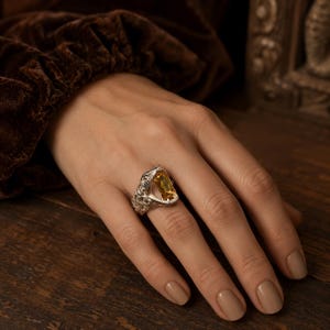 May include: A silver ring featuring an elongated rectangular yellow gemstone set in a textured, ornate metal band. The band displays intricate, raised scrollwork patterns across its surface, framing the central stone in a bold, open design.