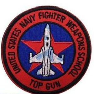 Pode incluir: Um emblema bordado circular com uma borda vermelha contendo o texto preto UNITED STATES NAVY FIGHTER WEAPONS SCHOOL e TOP GUN. O centro exibe um círculo azul com uma estrela vermelha atrás de uma silhueta branca de avião de caça.