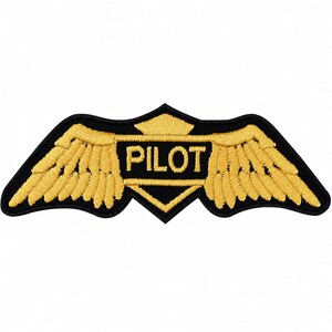 May include: An embroidered patch featuring a central black shield shape with the word PILOT stitched in yellow thread. The shield is flanked by two stylized yellow wings with black outlines, creating a wide, symmetrical emblem design.