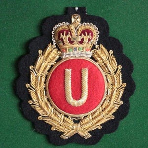 Puede incluir: Un parche bordado de estilo militar con un círculo rojo central que contiene una letra U dorada, rodeado por una corona de laurel dorada y rematado con una corona detallada. El emblema está sobre un fondo de fieltro negro con bordes festoneados.