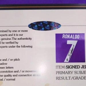May include: A rectangular certificate of authenticity with a black border on a white background. It features a blue holographic seal, text detailing STARSAUTHENTIC examination criteria, and a purple square with the white text RONALDO 7.