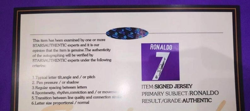May include: A rectangular certificate of authenticity with a black border on a white background. It features a blue holographic seal, text detailing STARSAUTHENTIC examination criteria, and a purple square with the white text RONALDO 7.
