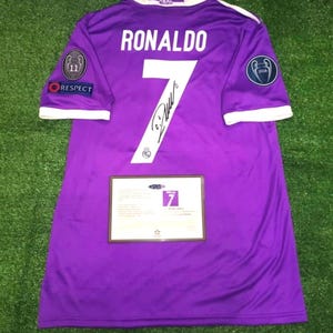 May include: A purple Real Madrid soccer jersey featuring the name RONALDO and the number 7 in white on the back. The jersey includes a black signature on the number, sleeve patches, and a rectangular certificate of authenticity card attached.