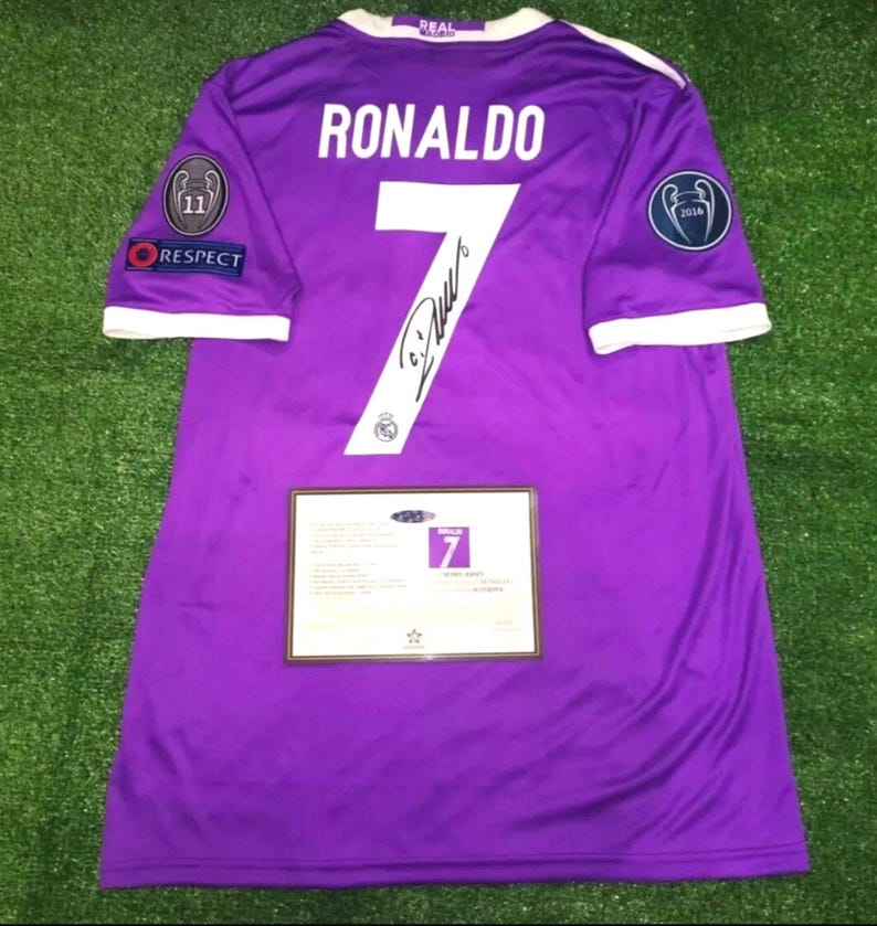 May include: A purple Real Madrid soccer jersey featuring the name RONALDO and the number 7 in white on the back. The jersey includes a black signature on the number, sleeve patches, and a rectangular certificate of authenticity card attached.