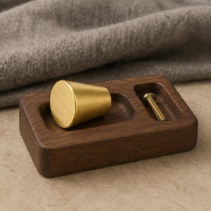 May include: A conical brass cabinet knob with a brushed finish resting on a rectangular dark brown wooden block with a recessed slot containing a single metal screw. The knob has a flat circular top and a tapered body with a smooth metallic surface.