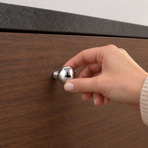 May include: A polished chrome spherical cabinet knob attached to a dark brown wood-grain drawer front. The knob features a smooth, reflective, round surface and a small cylindrical base that connects directly to the flat wooden panel surface.