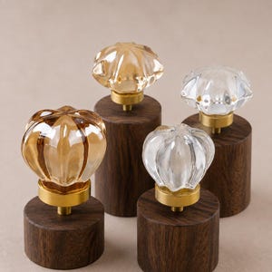 May include: Four cabinet knobs featuring fluted, pumpkin-shaped glass tops in clear and amber tones. Each glass piece is mounted on a circular brass-toned metal base, which is attached to a cylindrical dark brown wooden block base.