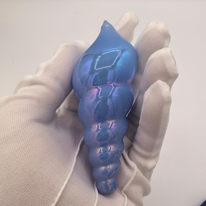 Könnte beinhalten: Ein spiralförmiges Glasornament mit einer glatten, irisierenden hellblauen Oberfläche. Das Objekt hat eine spitz zulaufende Oberseite und eine Reihe von abgerundeten, segmentierten Rippen, die zur Basis hin allmählich breiter werden.