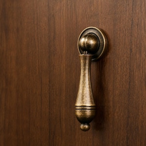 May include: A drop-style cabinet pull with an antique brass finish. The hardware features a circular mounting plate and a teardrop-shaped handle with horizontal grooved detailing near the base, attached to a rounded, ball-shaped pivot point.