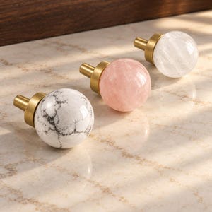 May include: Three spherical cabinet knobs featuring a brass-toned metal base. One knob is white with grey veining, one is solid pale pink, and one is translucent white. Each knob has a smooth, rounded surface and a short cylindrical mounting post.