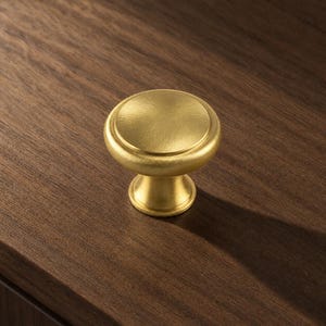 May include: A circular cabinet knob with a brushed gold finish. The knob features a flat, smooth top surface with a slightly rounded edge and a tapered, cylindrical base that connects to the mounting point, creating a uniform metallic appearance.