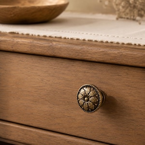 May include: A round, antique brass-toned metal drawer knob featuring a detailed, embossed floral leaf pattern with a beaded outer edge, mounted on the front of a light brown wooden drawer with a visible horizontal wood grain texture.