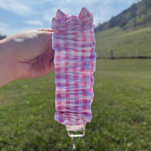 May include: A ruffled fabric keychain wristlet featuring a pink and white gingham check pattern. The fabric is gathered into a series of soft folds and is attached to a silver-toned metal hardware clasp with a circular split ring at the bottom.