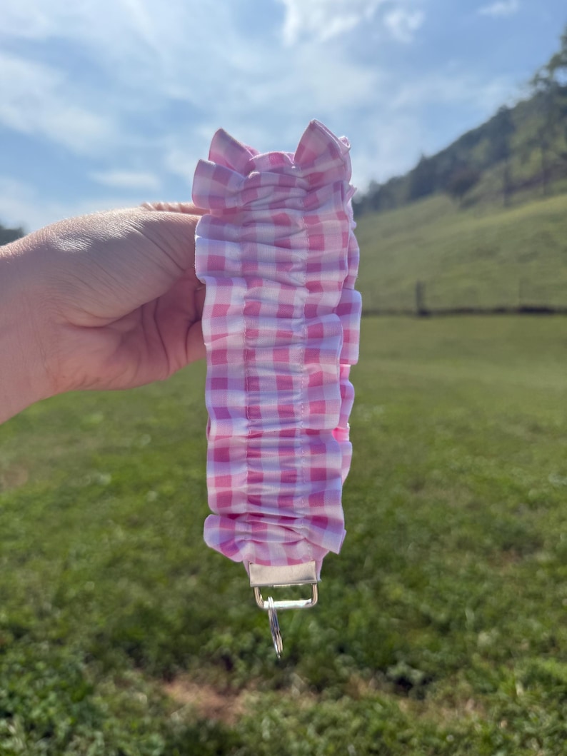 May include: A ruffled fabric keychain wristlet featuring a pink and white gingham check pattern. The fabric is gathered into a series of soft folds and is attached to a silver-toned metal hardware clasp with a circular split ring at the bottom.