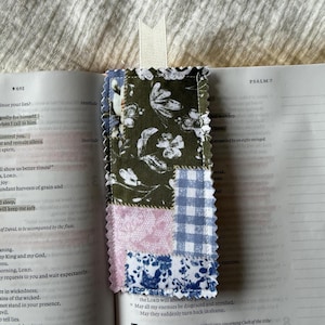 May include: A rectangular fabric bookmark featuring a patchwork design of olive green, light pink, and blue floral and gingham patterns with pinked edges and a small cream-colored ribbon tab at the top, placed inside an open book.