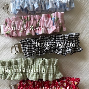 May include: Five fabric keychains with metal rings, each featuring a ruffled design. Patterns include blue floral, pink with berries and bows, black gingham, green gingham, and red floral with white daisies, labeled with their respective names.