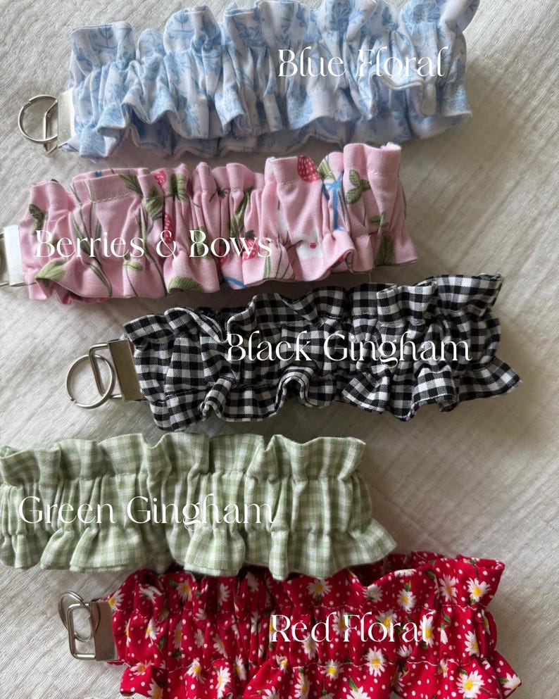 May include: Five fabric keychains with metal rings, each featuring a ruffled design. Patterns include blue floral, pink with berries and bows, black gingham, green gingham, and red floral with white daisies, labeled with their respective names.