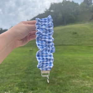 May include: A fabric wristlet keychain featuring a blue and white gingham checkered pattern. The fabric is gathered into a ruffled, scrunchie-style design and is attached to a silver-toned metal hardware clasp with a circular split ring attached.