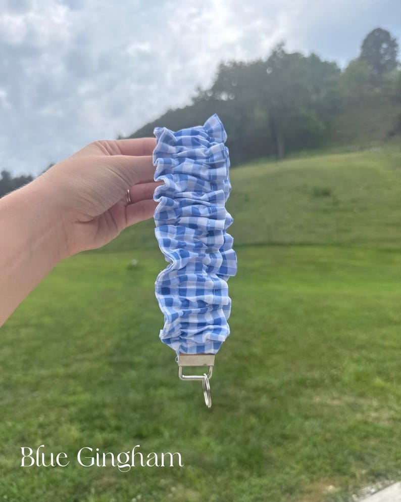 May include: A fabric wristlet keychain featuring a blue and white gingham checkered pattern. The fabric is gathered into a ruffled, scrunchie-style design and is attached to a silver-toned metal hardware clasp with a circular split ring attached.