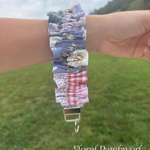 May include: A fabric wristlet keychain featuring a gathered, ruffled design with a patchwork pattern of blue, white, and red floral and gingham prints. The strap is attached to a silver-toned metal clasp with a circular split ring for keys.