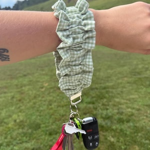 May include: A light green and white gingham fabric wristlet keychain featuring a ruffled, scrunchie-style design. The fabric band is attached to a silver-toned metal clasp holding a collection of metal keys, a black car key fob, and a small red flashlight.