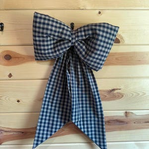 May include: A large decorative bow made of fabric with a navy blue and cream plaid pattern. The bow features two wide, structured loops at the top and two long, pointed tails hanging downward, creating a classic and symmetrical appearance.