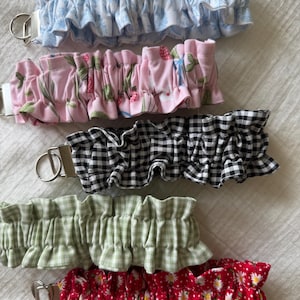 May include: A collection of five fabric wristlet keychains featuring ruffled designs. The patterns include light blue floral, pink strawberry, black and white gingham, sage green gingham, and red with white daisies. Each has a silver metal hardware ring.