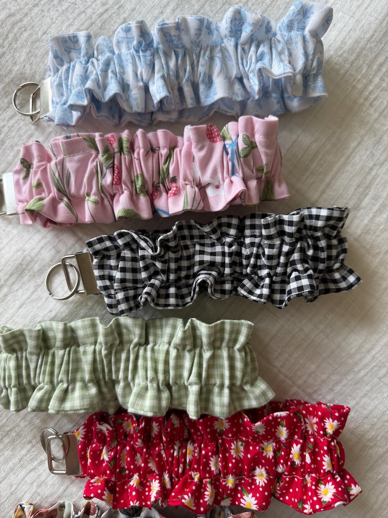 May include: A collection of five fabric wristlet keychains featuring ruffled designs. The patterns include light blue floral, pink strawberry, black and white gingham, sage green gingham, and red with white daisies. Each has a silver metal hardware ring.