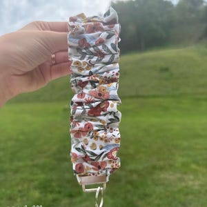 May include: A fabric wristlet keychain featuring a white background with a repeating pattern of red, orange, and yellow flowers, green leaves, and blue accents. The fabric is gathered into ruffles and attached to a silver metal clasp with a ring.