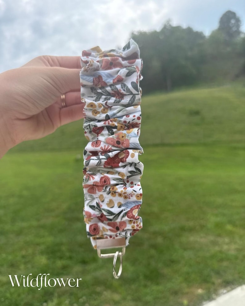 May include: A fabric wristlet keychain featuring a white background with a repeating pattern of red, orange, and yellow flowers, green leaves, and blue accents. The fabric is gathered into ruffles and attached to a silver metal clasp with a ring.