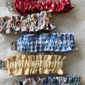 May include: A collection of five fabric wristlet keychains featuring ruffled designs. The patterns include Red Floral, Wildflower, Blue Gingham, Butter Yellow with a white grid, and Floral Patchwork. Each keychain has a silver metal hardware clasp.