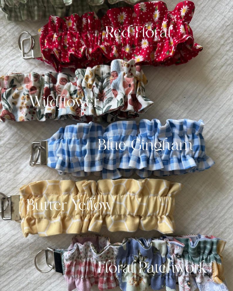 May include: A collection of five fabric wristlet keychains featuring ruffled designs. The patterns include Red Floral, Wildflower, Blue Gingham, Butter Yellow with a white grid, and Floral Patchwork. Each keychain has a silver metal hardware clasp.