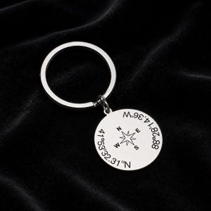 May include: A silver-coloured metal keyring featuring a circular pendant. The pendant is engraved with a central compass rose design and the coordinates 41°53'32.31"N and 88°28'14.36"W. The keyring includes a standard round split ring for keys.