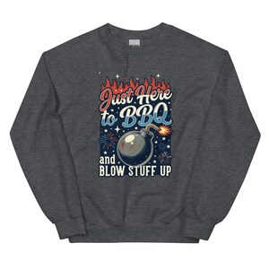 May include: A dark heather gray crewneck sweatshirt featuring a graphic print with red flames, blue fireworks, and a black bomb with a lit fuse. The text reads Just Here to BBQ and BLOW STUFF UP in a mix of red, white, and blue stylized lettering.