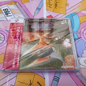 Puede incluir: Un videojuego Gradius II Gofer no Yabou para el sistema PC Engine Super CD-ROM. La caja del juego presenta una nave espacial plateada volando sobre un paisaje ardiente con el texto Gradius II, Gofer no Yabou, Super CD-ROM y Konami impreso.