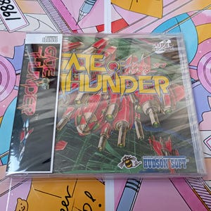 Può includere: Un videogioco Gate of Thunder sigillato per il sistema Super CD-ROM, con un'illustrazione di un'astronave rossa sulla copertina, il logo Hudson Soft e un'etichetta laterale verticale con il titolo in grassetto giallo e rosso su sfondo nero.