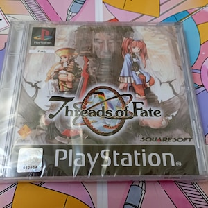Pode incluir: Uma caixa de jogo de vídeo PlayStation para Threads of Fate, com personagens de estilo anime, um emblema circular central e o logótipo da Squaresoft. A caixa de plástico transparente exibe a etiqueta da região PAL e um autocolante holográfico.