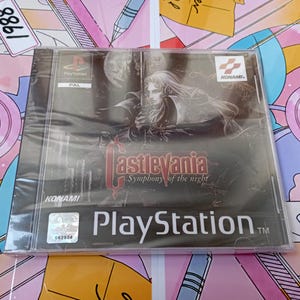 Może przedstawiać: Zafoliowana gra wideo Castlevania Symphony of the Night na konsolę PlayStation. Okładka przedstawia mroczną, gotycką ilustrację długowłosej postaci trzymającej miecz, z wyraźnie widocznym tytułem gry oraz logo firmy Konami na froncie.