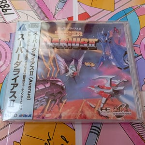 Puede incluir: Un videojuego Super Darius II para el sistema PC Engine CD-ROM en una caja de plástico transparente. La portada muestra naves espaciales y peces mecánicos coloridos sobre un fondo cósmico, con texto en japonés y el logotipo de Avenue.
