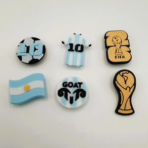 May include: A set of six shoe charms featuring soccer-themed designs: a black and white soccer ball with the number 10, a light blue and white striped jersey with the number 10, a gold FIFA trophy emblem, an Argentine flag, and a GOAT emblem.