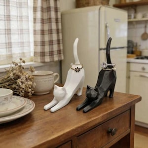 May include: Two cat-shaped ring holders, one in matte white and one in matte black, each featuring an elongated tail designed to hold rings. The white cat displays a gold-toned flower ring, while the black cat holds a silver-toned ring with a clear stone.