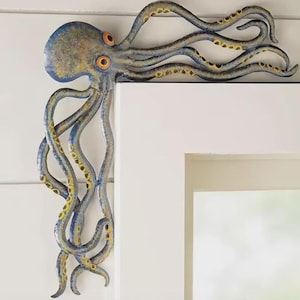 Op de afbeelding: Een blauwe en grijze metalen octopus wanddecoratie ontworpen om in een hoek te passen. De octopus heeft een textuurafwerking, opvallende oranje ogen en lange, gebogen tentakels met zichtbare gele zuignappen langs de randen.
