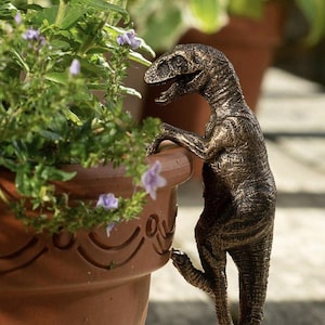 May include: A bronze-colored dinosaur figurine with a textured, weathered finish, depicted in a climbing pose with its front claws hooked over the rim of a terracotta-colored plant pot, featuring a detailed scaly skin texture and an open mouth.